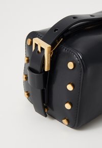 Borsa di pelle nera con borchie metalliche dorate, forma rettangolare e fibbia sicura. Presenta una superficie testurizzata e una tracolla regolabile.