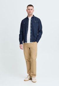 Dark blue button-up shirt with two chest pockets, paired with beige trousers and white sneakers with brown accents, standing against a plain background.