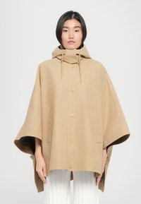 Poncho à capuche beige en tissu doux et texturé, doté de boutons-pression dorés, de poches latérales et de larges manches.