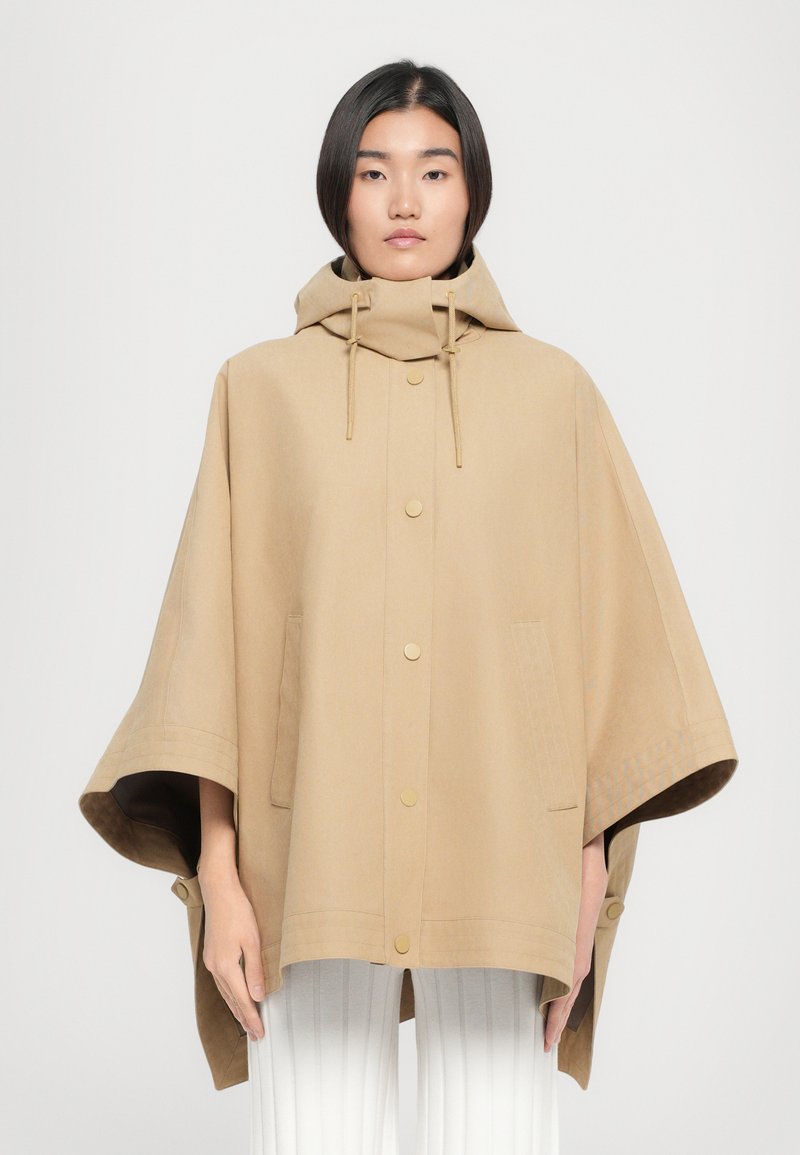 Poncho à capuche beige en tissu doux et texturé, doté de boutons-pression dorés, de poches latérales et de larges manches.