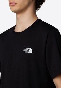 Ung person iført en sort The North Face T-shirt med et hvidt logo på venstre bryst, viser hals og delvist ansigt.