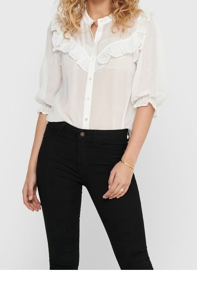 Chemise blanche à boutons avec des ruchés sur le devant et les manches, associée à un jean noir. Tissu transparent avec un motif en grille.