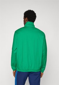 Moški s kratkimi kodrastimi lasmi, oblečen v svetlo zeleni jopič in modre hlače z modrimi črtami, stoji obrnjeno stran od bel background.