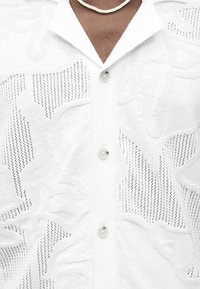 Κοντινό πλάνο ενός ατόμου που φοράει ένα λευκό πλεκτό πουκάμισο με γιακά, κουμπιά μπροστά και διακριτικό λουλουδένιο μοτίβο, συνοδευόμενο από λευκό κολιέ με χάντρες.