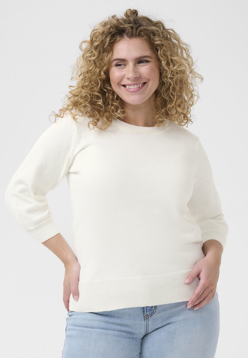 Cream-colored knit sweater with a round neckline and three-quarter sleeves, featuring a ribbed hem and soft texture. Worn with light blue jeans.