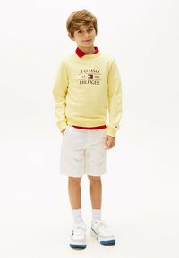 Sudadera amarilla con el logo "TOMMY HILFIGER", pantalones cortos blancos y zapatillas blancas con acentos azules. Un color rojo asoma de la sudadera.