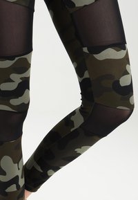 Kamouflage leggings med paneler av solid svart mesh och olivgröna mönster, tillverkade av elastiskt material med en slät textur.