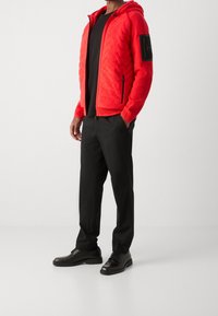 Man som bär en röd quiltad zip-up hoodie, svart crew neck-tröja, svarta byxor och svarta läder snörskor mot en enfärgad bakgrund.