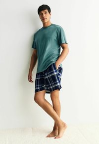 Vaaleanvihreä puuvillapaita yhdistettynä tummansinisiin ruutuhousuihin, joissa on valkoisia ja vaaleansinisiä yksityiskohtia. Malli seisoo paljain jaloin yksinkertaista taustaa vasten.