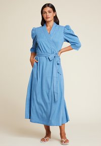Blå denim omslagkjole med korte puffermer, belte midje, sidelommer og en flytende midi-lengde silhuett.