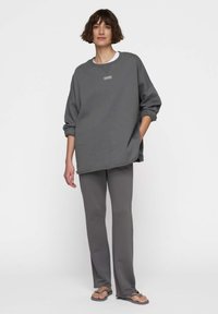 Felpa grigia over size con scollo rotondo e pantaloni coordinati grigi a zampa. Il tessuto appare liscio e morbido, con un delicato dettaglio del logo.
