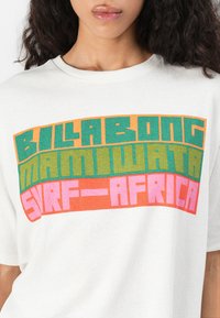 Sieviete, kura valkā baltu T-kreklu ar krāsainu bloku tekstu uz priekšu, kas lasāms "Billabong Mamuwata Surf-Africa".