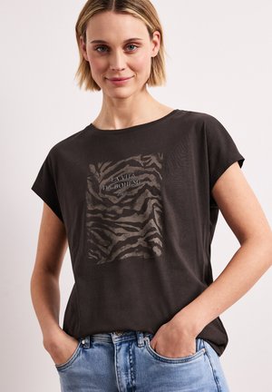 Ung kvinde med kort blondt hår iført sort t-shirt med tigerprint og lyseblå jeans, hænderne i lommerne, neutral baggrund.