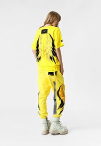 Conjunto de ropa amarilla que incluye una camiseta con gráficos de alas negras en la parte posterior y pantalones a juego. El conjunto incluye botas de combate blancas.