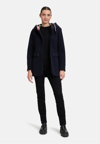 Marineblauer Wollmantel mit Kapuze, zwei Fronttaschen und einem bunten Karostofffutter. Kombiniert mit einem schwarzen Oberteil, schwarzen Hosen und schwarzen Stiefeln.