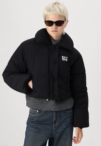 Sort cropped puffer-jakke med stor krave, trykknapper og hvid "EXTEND" logo. Har en grå strikket kant og sidelommer.