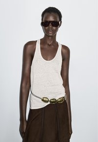 Vrouw met donkere zonnebril, witte mouwloze top, bruine rok en een riem met grote gouden ovale accenten tegen een effen lichte achtergrond.