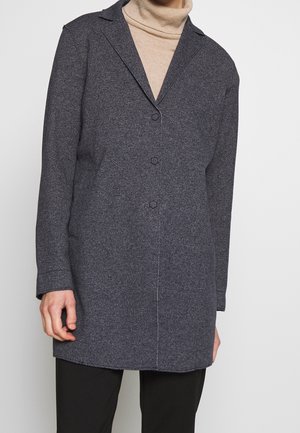 Personne portant un manteau gris foncé boutonné sur un pull beige à col roulé et un pantalon noir, debout devant un fond blanc uni.
