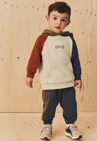 Dieťa oblečené v kapucňovej mikine s farebnými blokmi v zelených, hnedom a krémových paneloch s predným vreckom, kombinované s tmavomodrými nohavicami s olivovými pruhmi na bokoch.