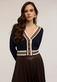 Mladá žena s dlouhými vlnitými vlasy má na sobě námořnický pletený cardigan s krémovými a hnědými pruhy, zlaté knoflíky, velké zlaté náušnice a tmavou plisovanou sukni.