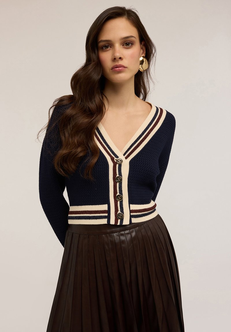Mladá žena s dlouhými vlnitými vlasy má na sobě námořnický pletený cardigan s krémovými a hnědými pruhy, zlaté knoflíky, velké zlaté náušnice a tmavou plisovanou sukni.