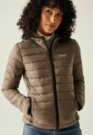 Mujer con cabello rizado que lleva una chaqueta acolchada taupe con cremallera negra y manos en los bolsillos, combinada con vaqueros azules y un jersey de cuello alto blanco.