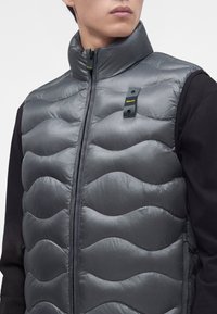 Homme portant un gilet sans manches matelassé gris par-dessus une chemise noire à manches longues, avec un col montant et une fermeture éclair frontale fermée.