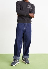 Černá mikina s bílým logem Adidas, spárovaná s širokými modrými džínami a šedými teniskami s bílými akcenty. Zelené pozadí.