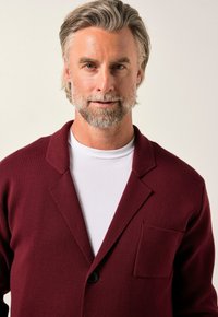 Burgundy stickad blazer med snedskurna slag, enkel knappstängning och en ficka på vänster sida. Bärs över en vit t-shirt.