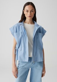  Lichtblauwe mouwloze jas met twee voorzakken, ritssluiting en koordjes. Draagt over een witte textuurtop en blauwe spijkerbroek.