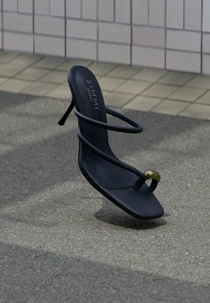 Μαύρο πέδιλο με λουράκια και τακούνι, με δαχτυλίδι στη μύτη που φέρει χρυσή χάντρα, να αιωρείται πάνω από υφασμένο πλακόστρωτο και φόντο με πλακάκια τοίχου.