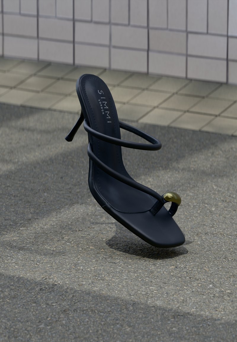 Μαύρο πέδιλο με λουράκια και τακούνι, με δαχτυλίδι στη μύτη που φέρει χρυσή χάντρα, να αιωρείται πάνω από υφασμένο πλακόστρωτο και φόντο με πλακάκια τοίχου.