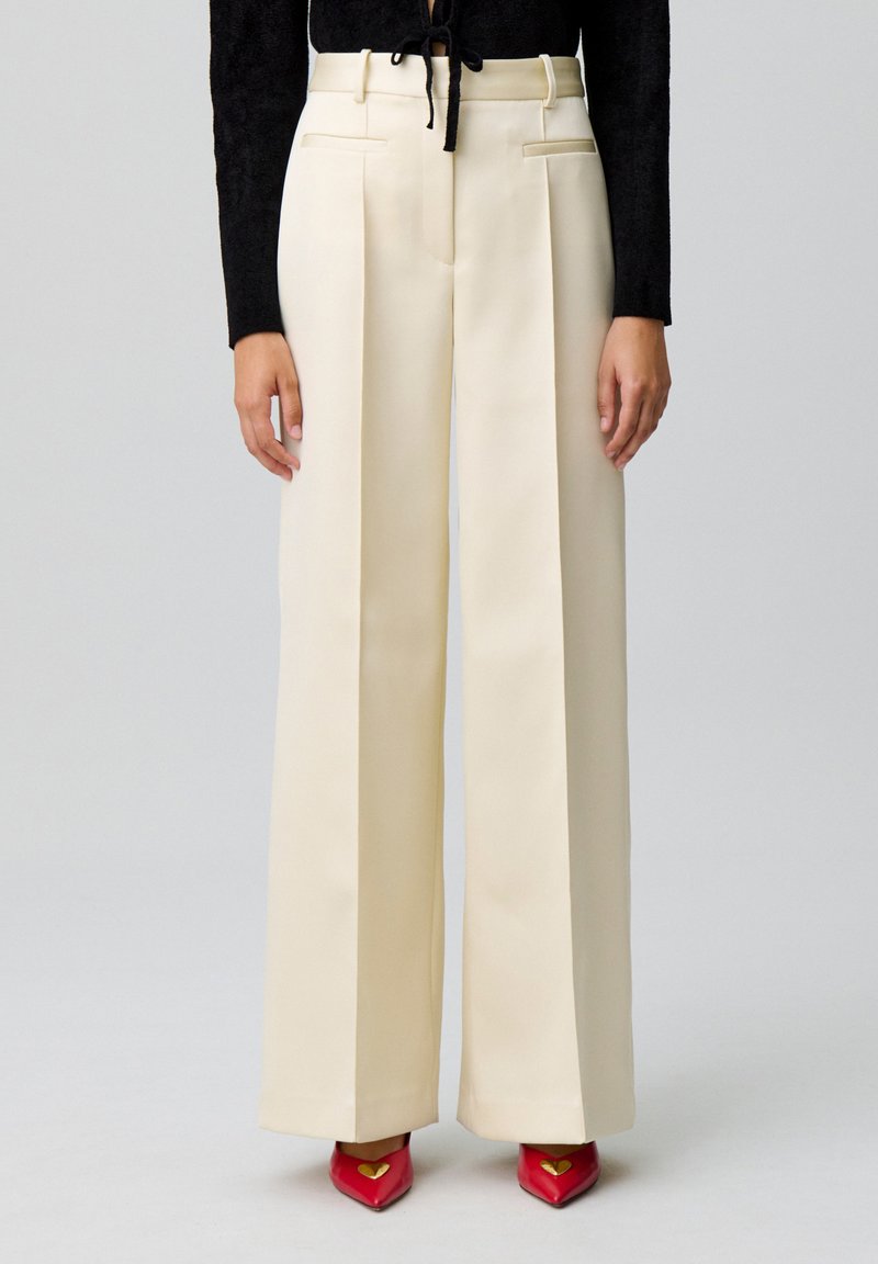 Pantalons larges taille haute crème avec plis à l'avant, associés à des chaussures rouges à bout pointu avec un accent en forme de cœur.