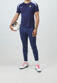 Námořnické sportovní tričko s modrými akcenty a logem, spárované s námořnickými legínami a bílými kopačkami s červenými detaily, držící vzorovaný fotbal.
