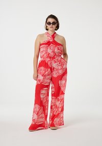 Rode jumpsuit met grote witte bloemenpatronen, mouwloos met een strikdetail bij de hals, wijde benen en een relaxte pasvorm. Draag het met zwarte zonnebrillen.