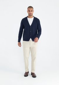 Marineblaues Strickblazer mit Revers, zwei Fronttaschen und drei Knöpfen; getragen über einem weißen Hemd und kombiniert mit beigen Hosen und braunen Schuhen.