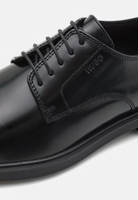 Chaussure habillée en cuir noir avec une finition brillante, design à lacets, accents cousus et semelle en caoutchouc avec un branding subtil.