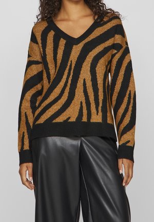 Femme portant un pull à col en V marron et noir à rayures zèbre avec manches longues et pantalon en cuir noir.