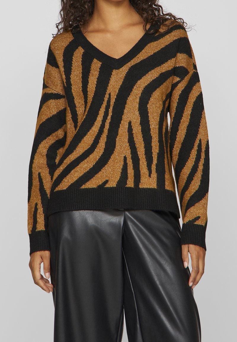 Femme portant un pull à col en V marron et noir à rayures zèbre avec manches longues et pantalon en cuir noir.