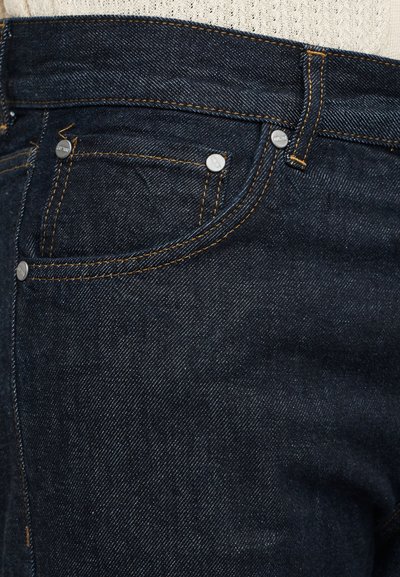 Μακροεντολή σκουρο μπλε τζιν με μπροστινή τσέπη που έχει πορτοκαλί ραφές και ασημένιες καρφίτσες πάνω σε ανοιχτόχρωμο υφασμάτινο φόντο.