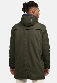 Man with dreadlocks wearing an olive green hooded jacket with black collar details, facing away against a plain light background.