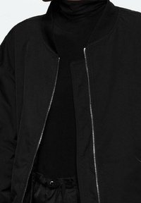 Zwarte bomberjack met een ritssluiting aan de voorkant, geribbelde kraag en lichtgewicht stof; gedragen over een zwarte turtleneck, die een gestroomlijnd ontwerp laat zien.