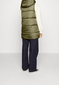 Gilet imbottito verde oliva con cappuccio, caratterizzato da sezioni trapuntate orizzontali. Abbinato a jeans in denim scuri a vita alta e sneakers nere.
