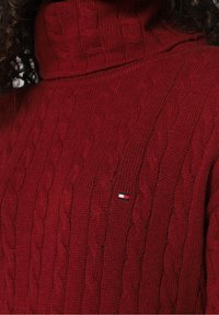 Maglione a collo alto lavorato a maglia rossa con un motivo testurizzato e una piccola toppa con logo sul lato sinistro. Materiale in maglia morbido e spesso.