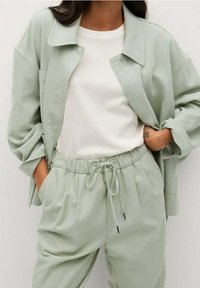 Světle zelená, volně střižená bunda s velkým límcem a patentkami, kombinovaná se sladěnými kalhotami s elastickým pasem a šňůrkou. Bílé tričko pod tím.