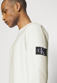 Hanorac alb cu guler rotund, dotat cu manșete ribbed și o etichetă din material negru pe mâneca stângă cu logo-ul "Calvin Klein Jeans" în alb.