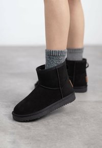 Botines de ante negro con punta redonda y suela de goma texturizada. Llevados con calcetines de rayas grises, que presentan un sutil detalle de logo en el talón.