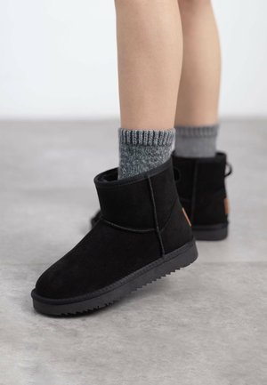 Botines de ante negro con punta redonda y suela de goma texturizada. Llevados con calcetines de rayas grises, que presentan un sutil detalle de logo en el talón.