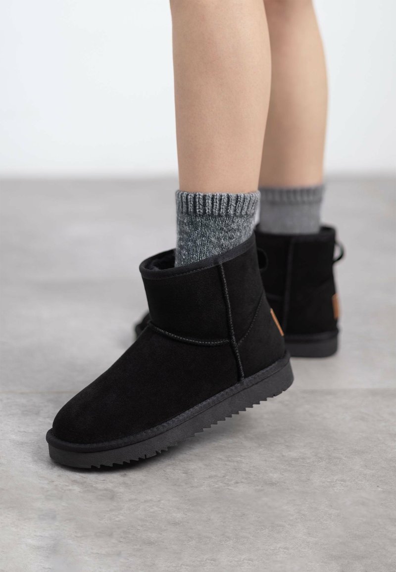 Botines de ante negro con punta redonda y suela de goma texturizada. Llevados con calcetines de rayas grises, que presentan un sutil detalle de logo en el talón.