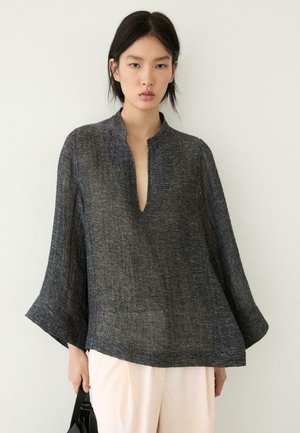 Jonge vrouw die een losse donkergrijze lange mouwtop met structuur en diepe halslijn draagt, gecombineerd met lichtbeige broek, en een zwarte handtas vasthoudt tegen een effen achtergrond.